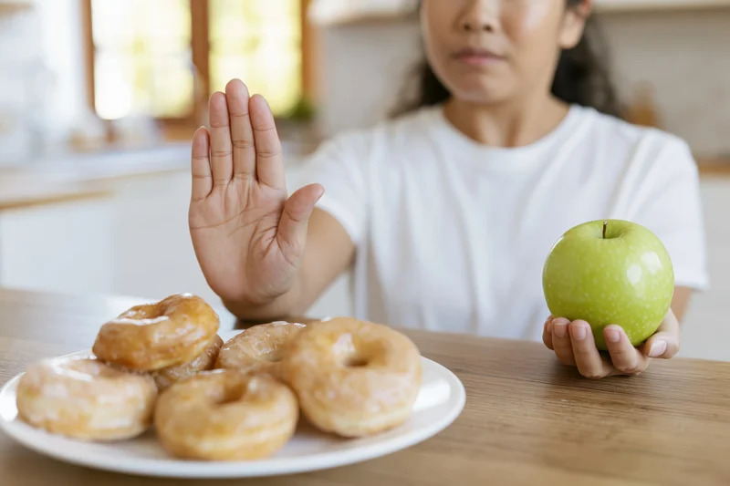 Asian woman holding green apple and pushing away plate of glazed donuts at kitchen table