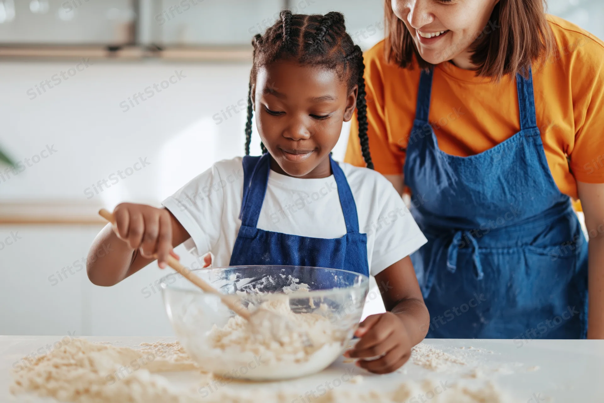 Young black girl mixing flour in a bowl with wooden spoon while woman smiles beside her in kitchen