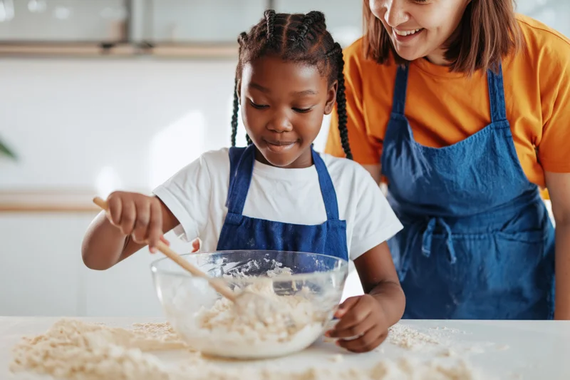 Young black girl mixing flour in a bowl with wooden spoon while woman smiles beside her in kitchen