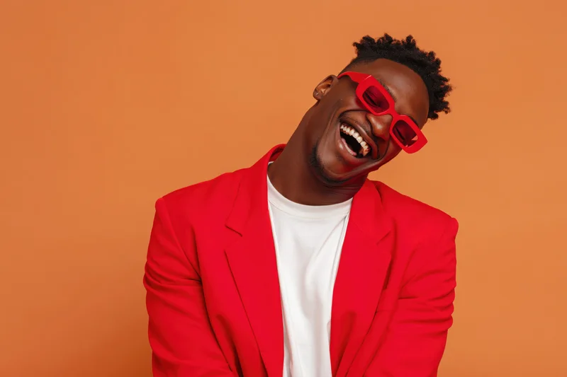 Young black man laughing with red sunglasses and red blazer over white t-shirt against orange background