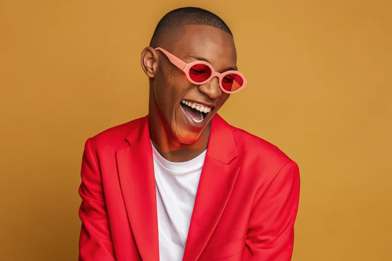 Young black man laughing in red suit and pink sunglasses against orange background