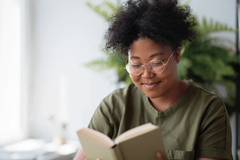 Young black woman with curly hair and round glasses reading a book indoors smiling