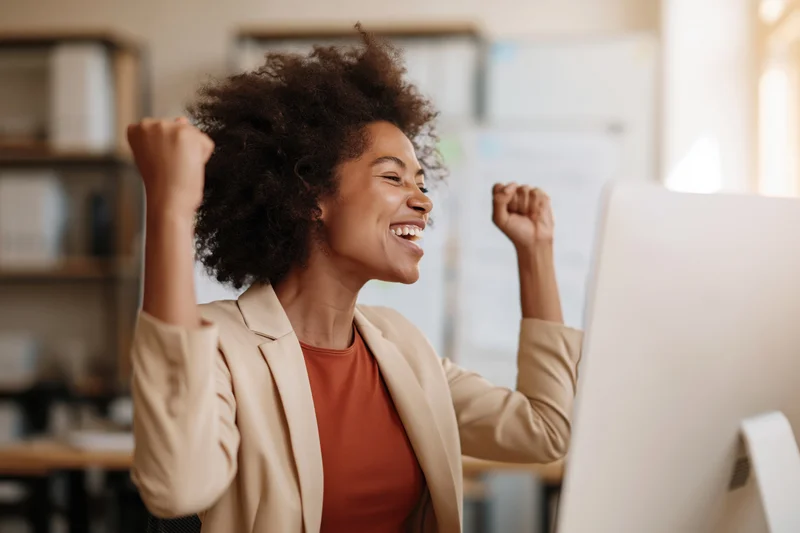 Young black woman celebrating success at office computer with fists raised