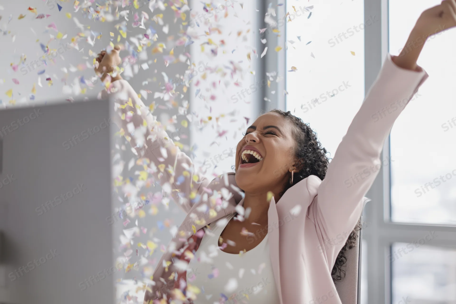 Woman laughing with arms raised amidst falling confetti at office desk