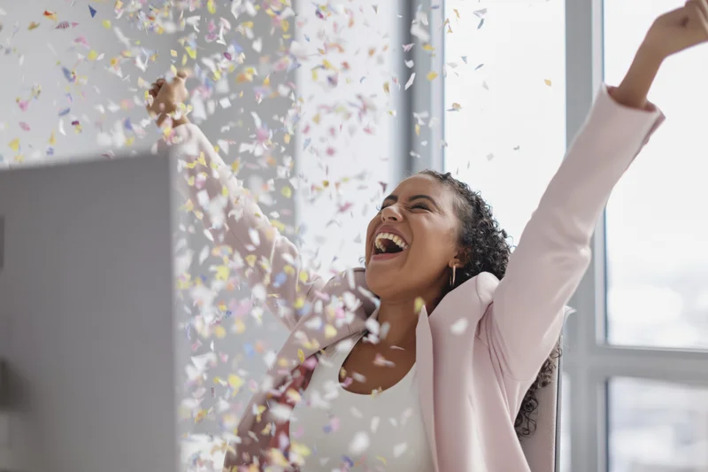 Woman laughing with arms raised amidst falling confetti at office desk