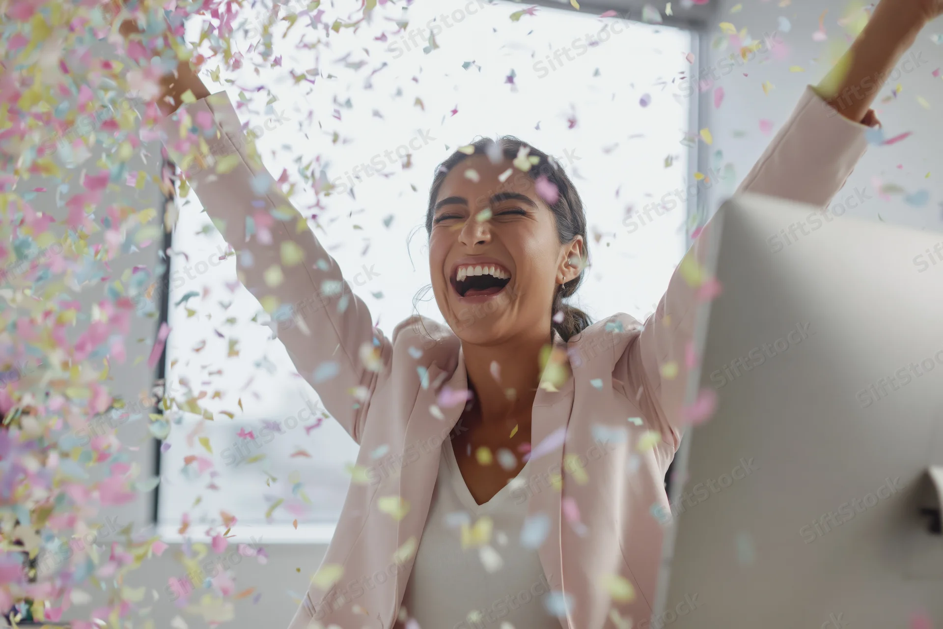 Woman laughing with arms raised amidst falling confetti at office desk