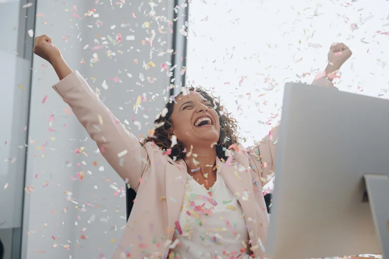 Woman celebrating success at office desk with confetti falling around her