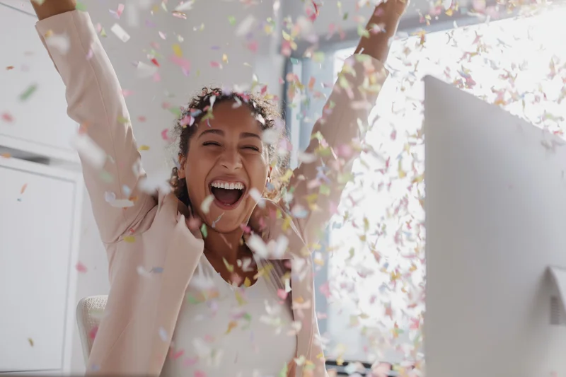 Woman laughing with arms raised amid falling confetti at office desk