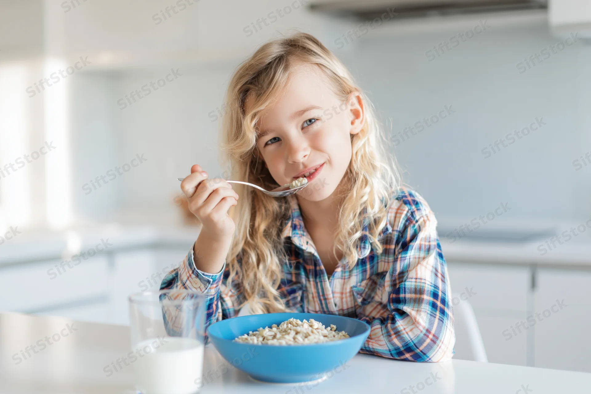 Young blonde girl eating cereal from a blue bowl at a kitchen table
