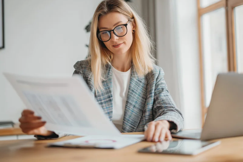 Woman in plaid blazer reviewing documents at office desk with laptop and smartphone nearby