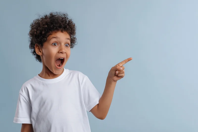 Young boy with curly dark hair pointing excitedly while wearing a white tshirt against a light blue background