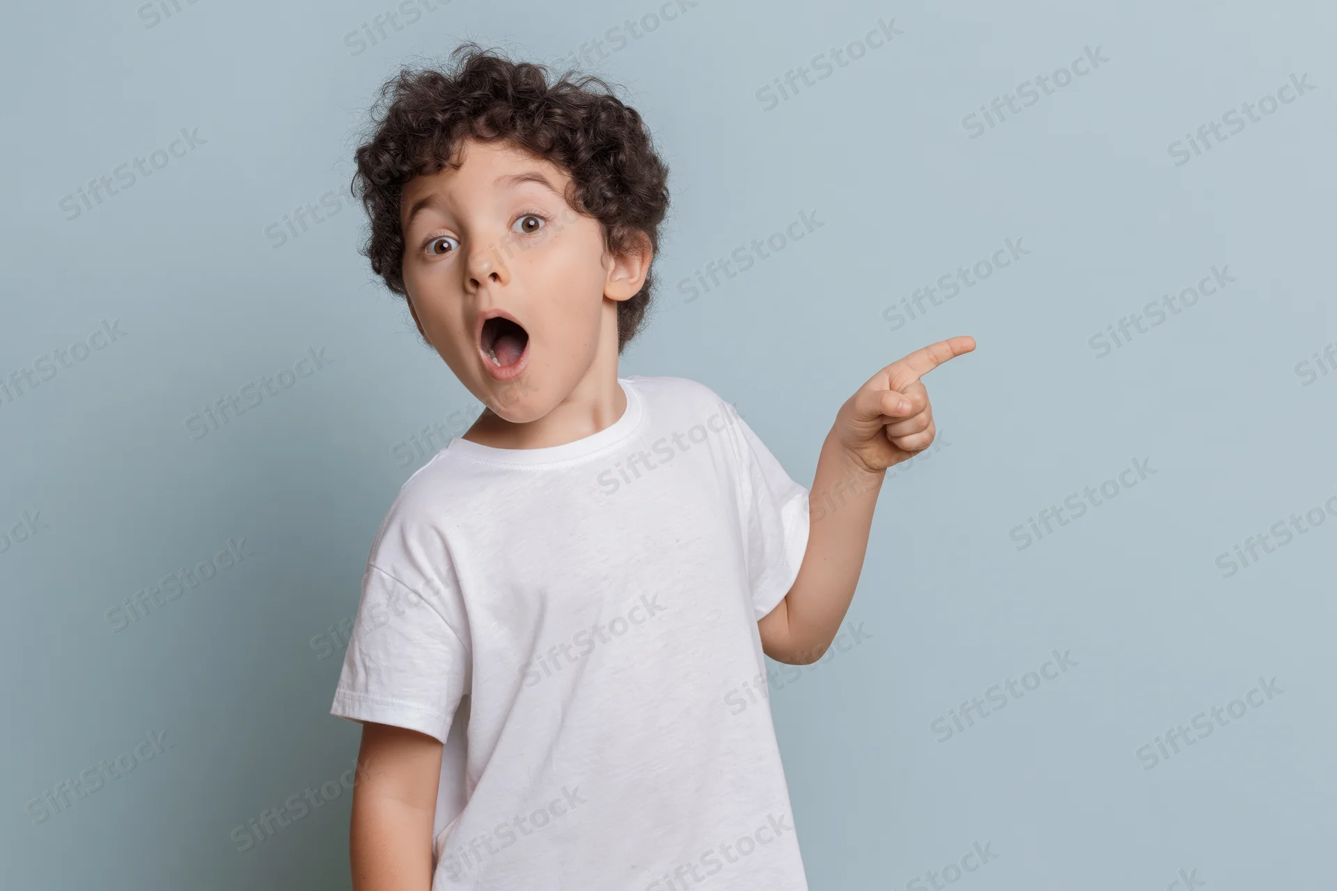 Young boy with curly dark hair pointing and wide open mouth in surprise against light blue background