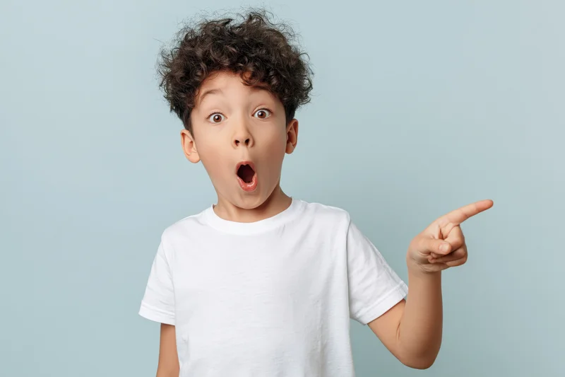 Young boy with curly hair pointing and wide open mouth in surprise against light blue background