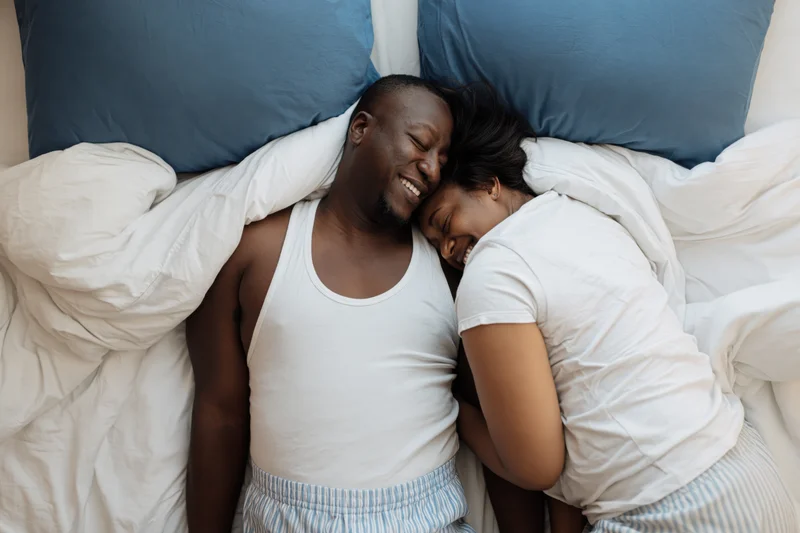 Young african couple laughing and cuddling in bed together, wrapped in white sheets with blue pillows
