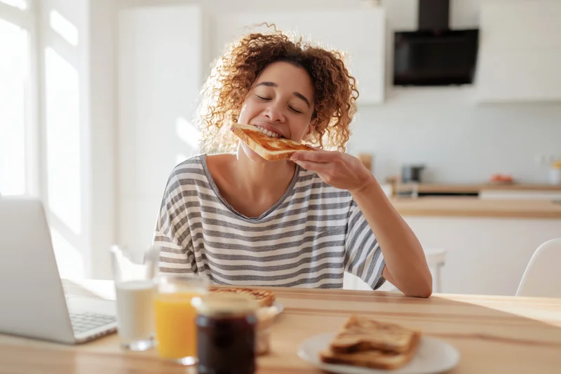 Woman biting into toast at kitchen table with laptop and breakfast items