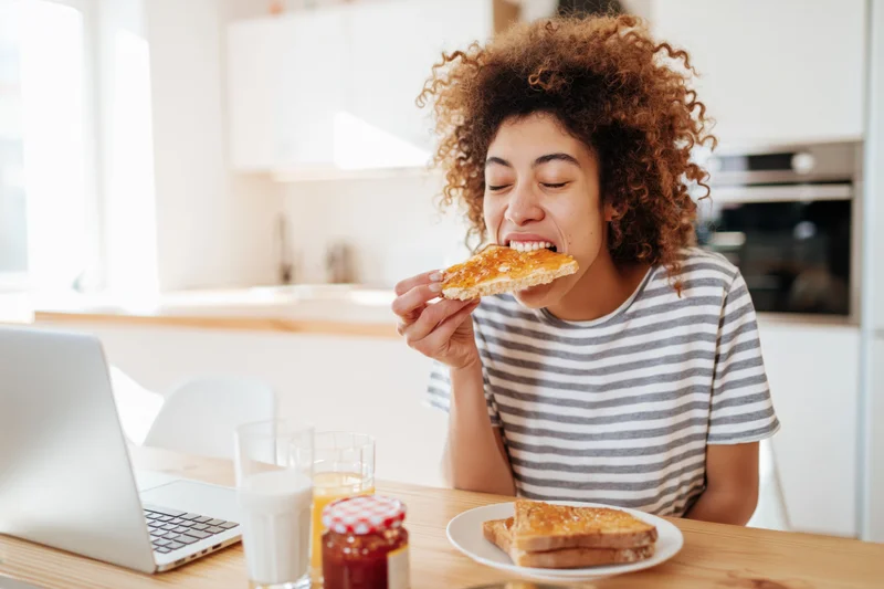 Woman biting into toast at kitchen table with laptop and breakfast items