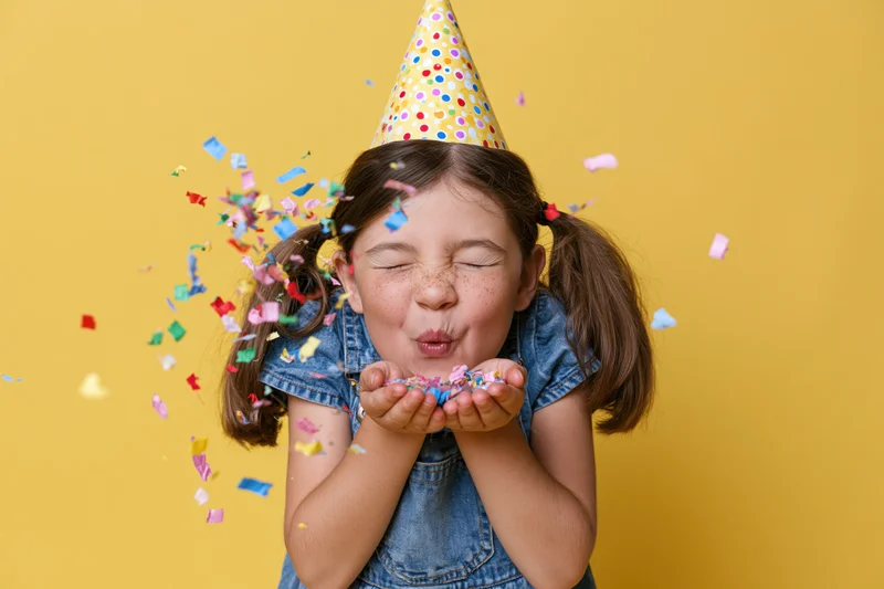 Young girl blowing colorful confetti from her hands while wearing a party hat and denim overalls against a yellow background