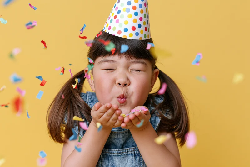 Young girl with pigtails blowing colorful confetti while wearing a party hat against a yellow background