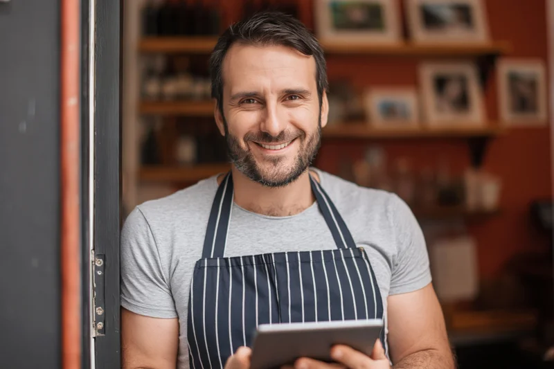 Smiling man in striped apron holding tablet at cafe doorway