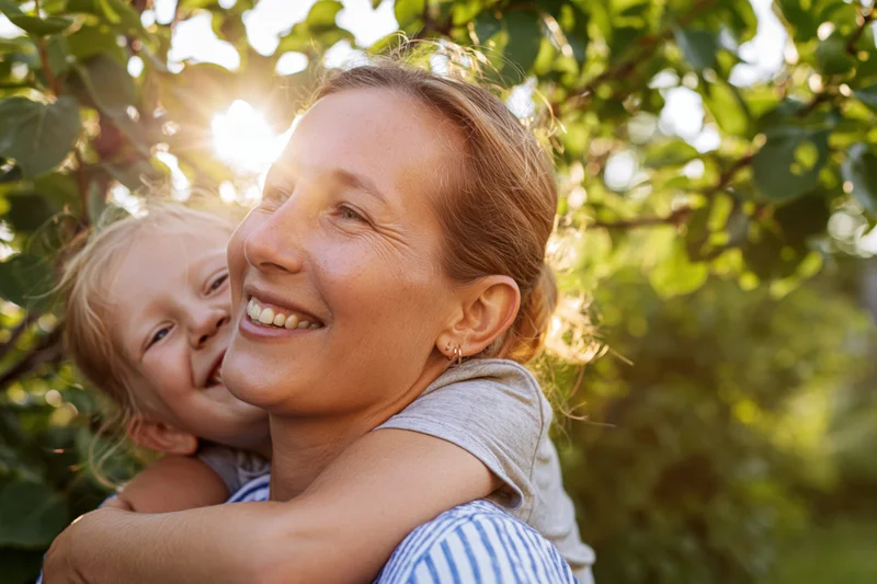 Mother carrying daughter on her back in a sunlit garden