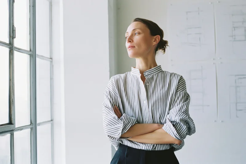 Woman architect standing by window with arms crossed in modern office studio