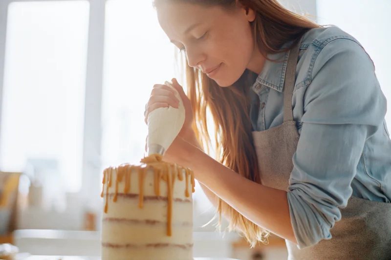 Woman decorating a layered cake with caramel icing in a bright kitchen
