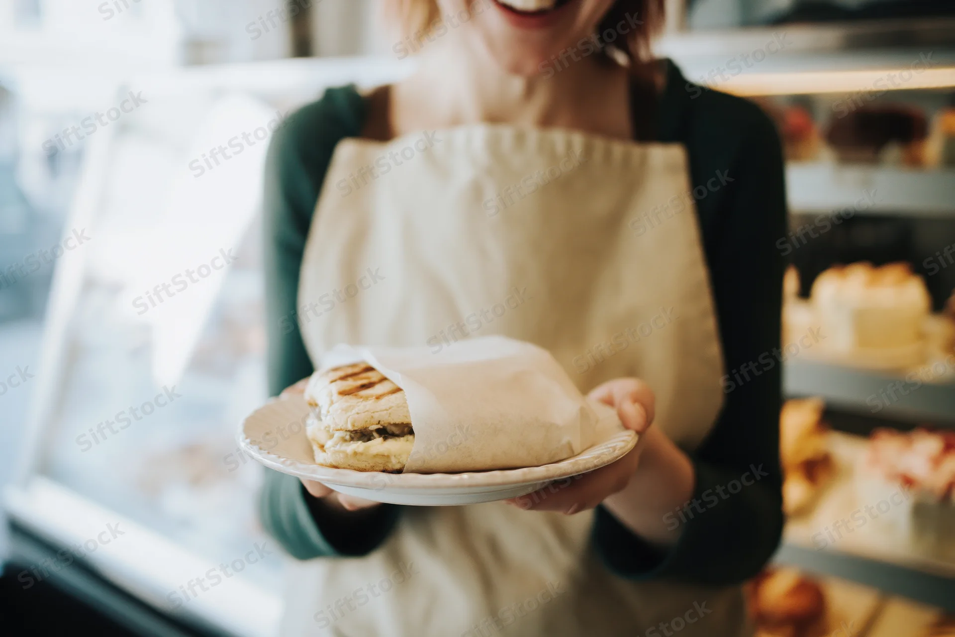 Smiling woman in apron holding plate with sandwich in cafe bakery setting