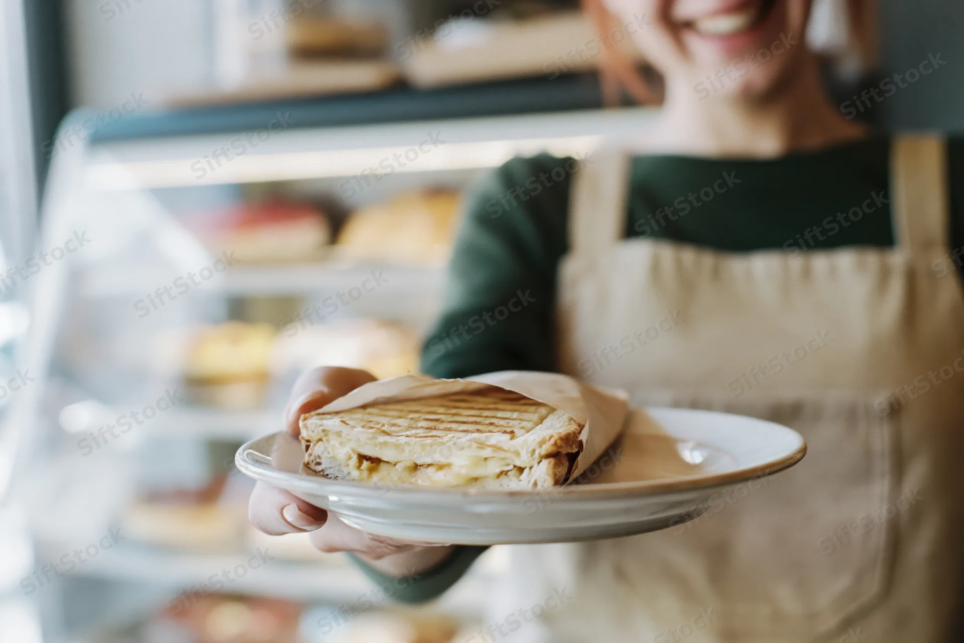 Smiling woman in apron holding plate with sandwich in cafe interior