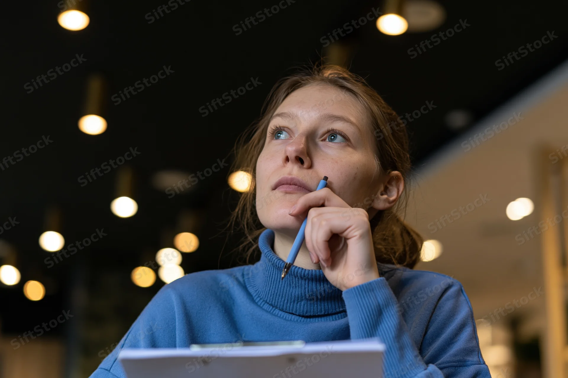 Woman holding pencil to chin while looking up, seated in modern cafe lounge with warm pendant lights