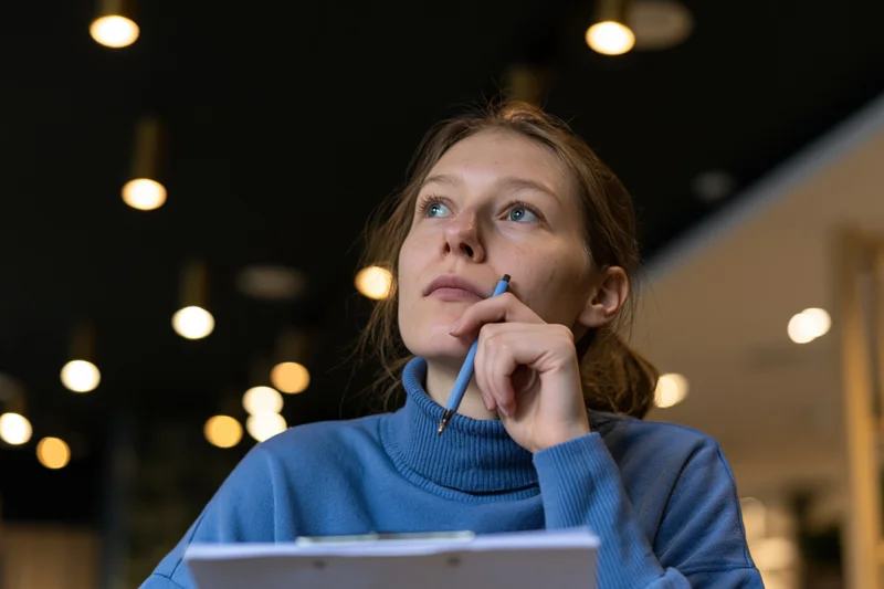 Woman holding pencil to chin while looking up, seated in modern cafe lounge with warm pendant lights