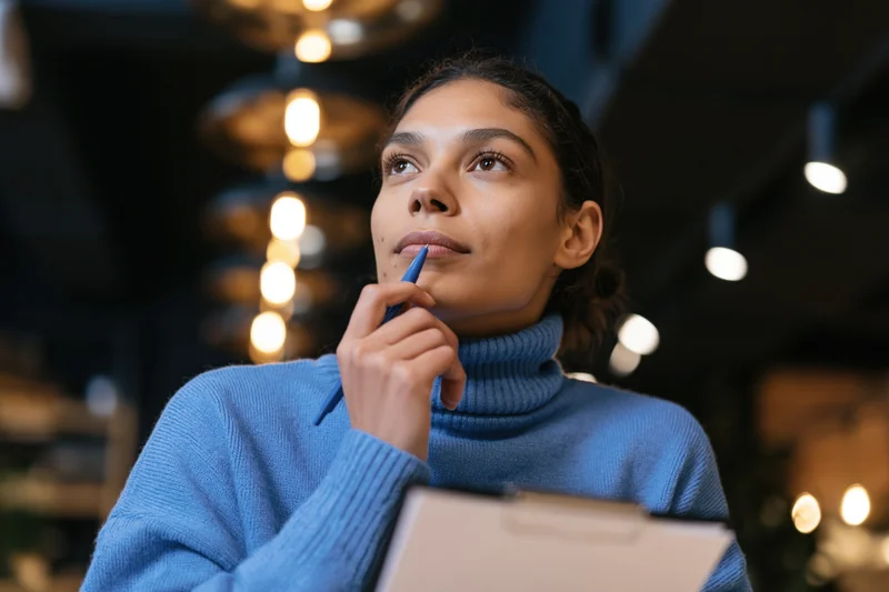 Woman thinking with pen to chin holding clipboard in modern cafe lounge