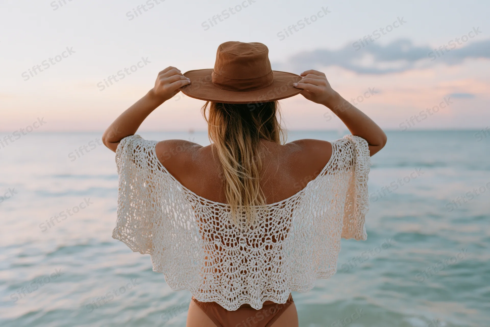 Woman from behind holding wide-brim hat on beach at sunset