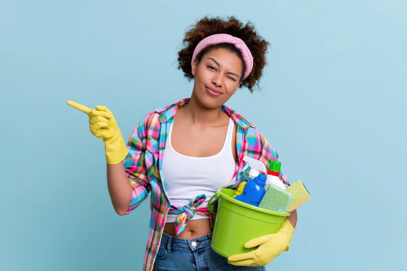 Woman winking and pointing while holding a green bucket of cleaning supplies against a blue background
