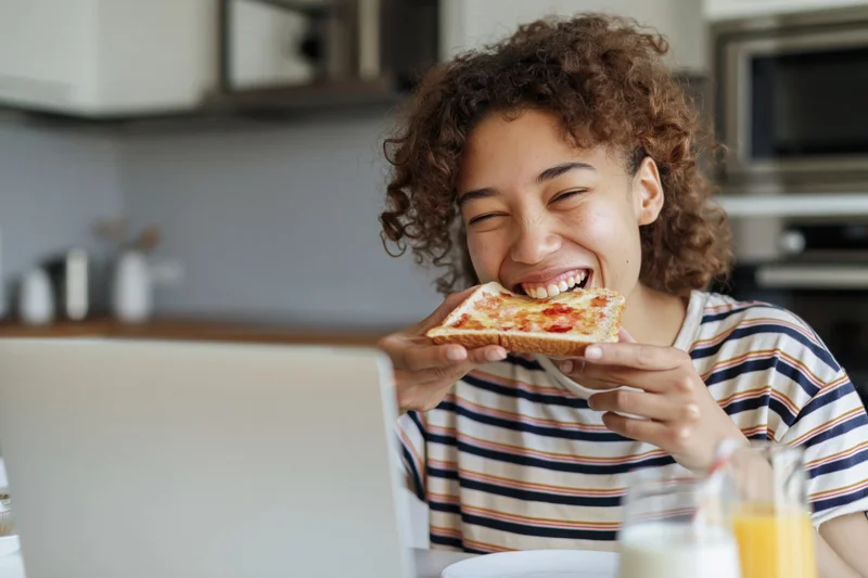 Woman eating toast with jam while smiling at laptop in kitchen