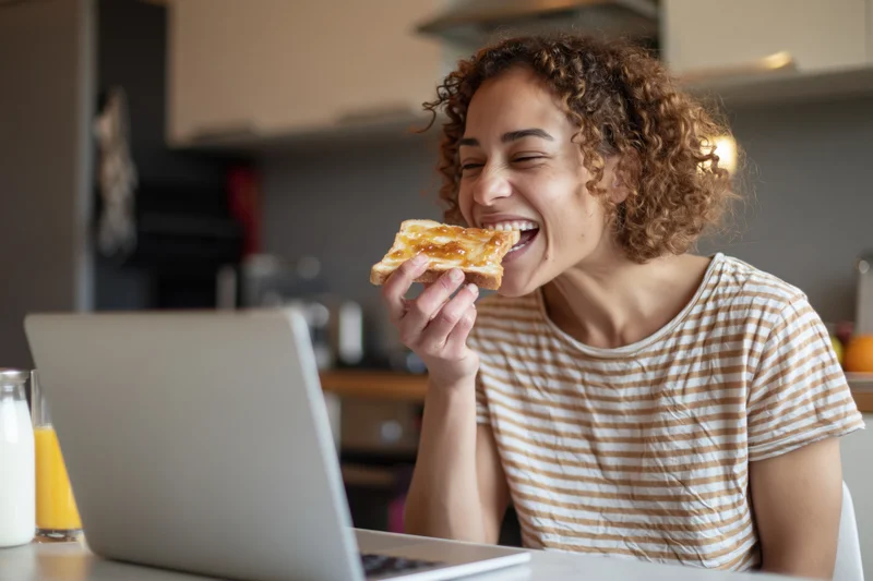 Woman laughing while eating toast with jam at a kitchen table in front of a laptop