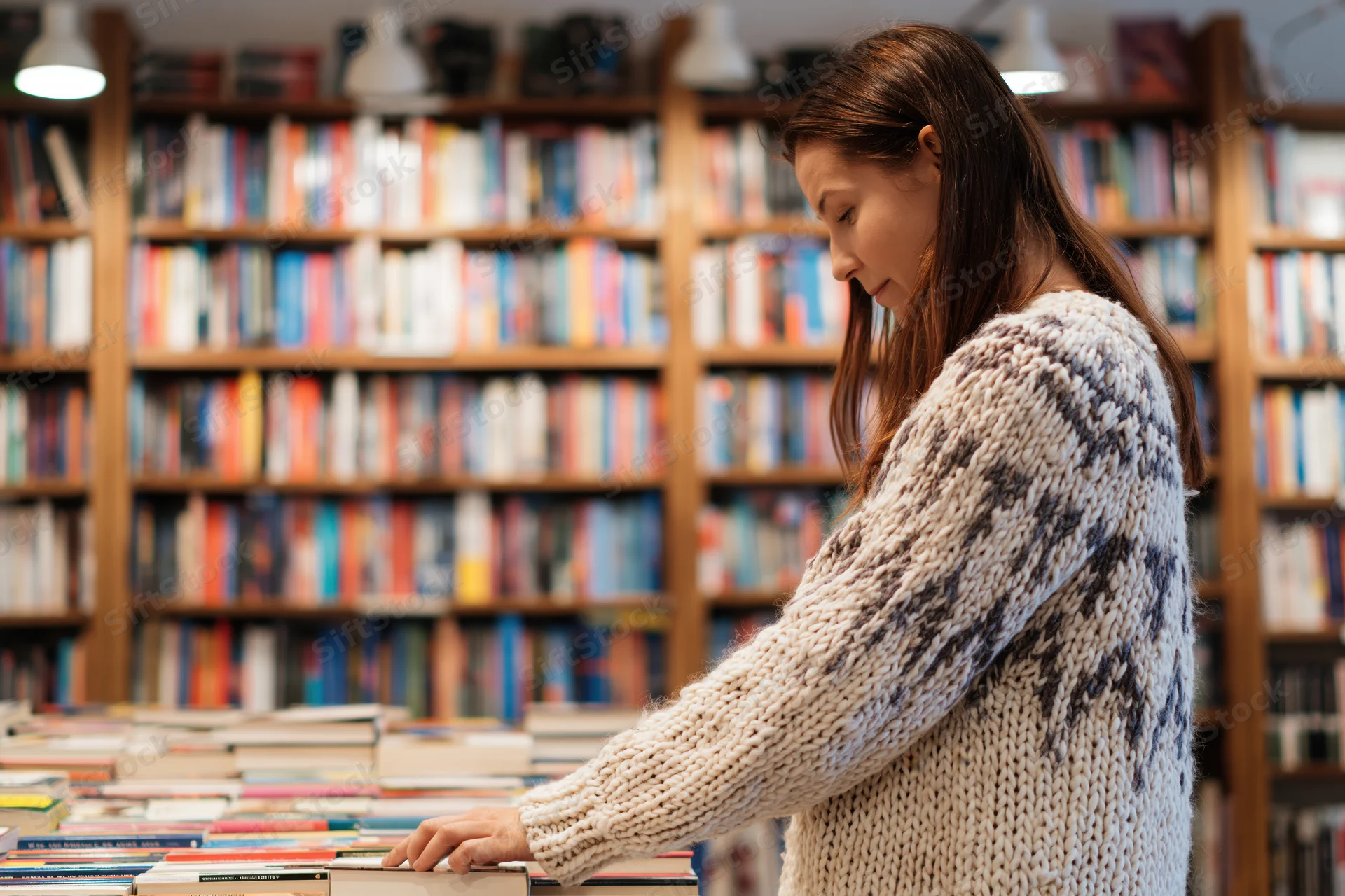 Woman browsing books in a bookstore, wearing a cream knit sweater, surrounded by wooden shelves filled with books