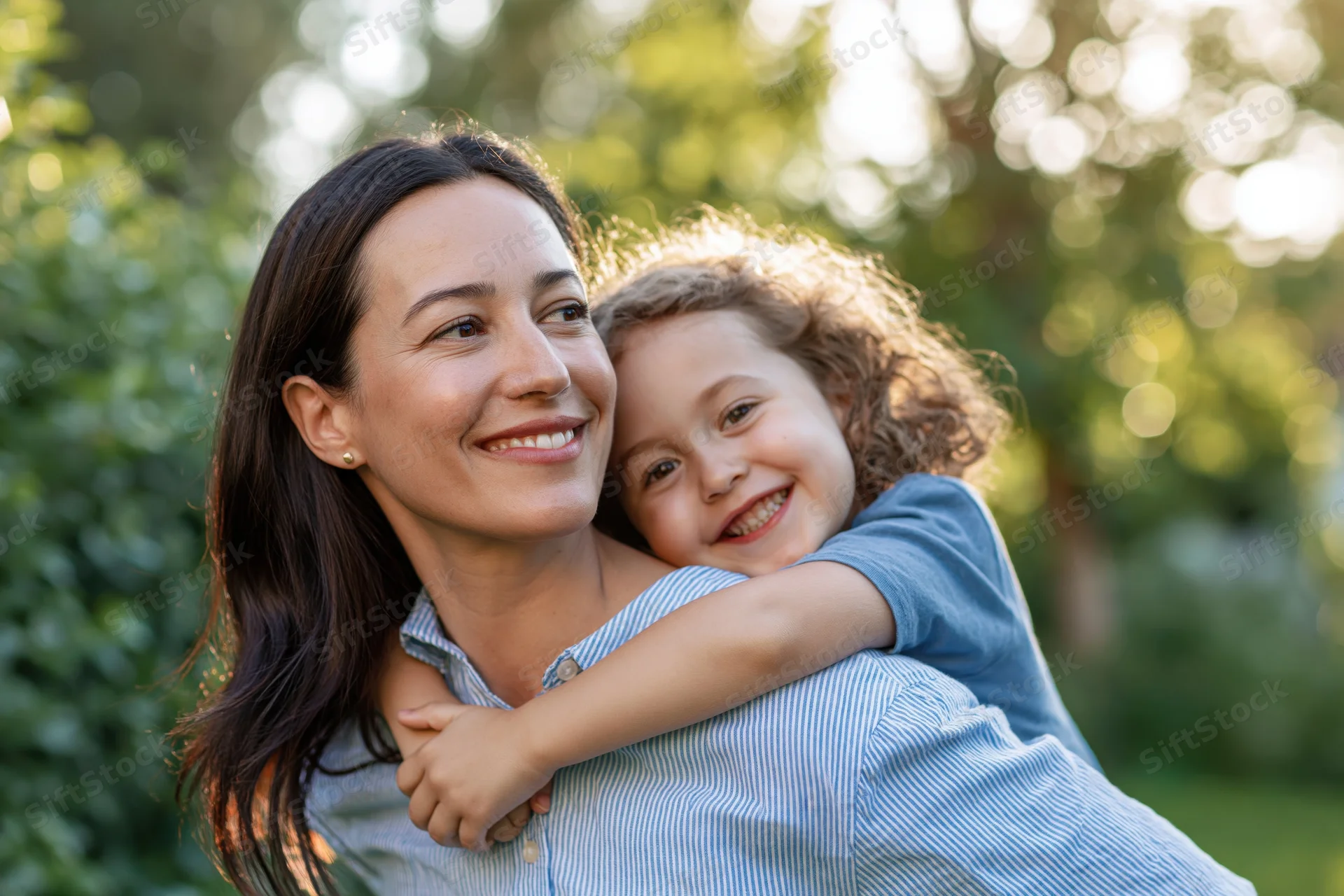 Woman carrying child on her back in a sunlit garden, both smiling and embracing
