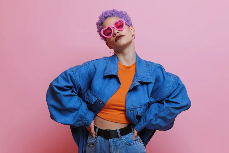 Young woman with short curly purple hair and pink heart-shaped sunglasses posing confidently in denim jacket and orange crop top against pink background