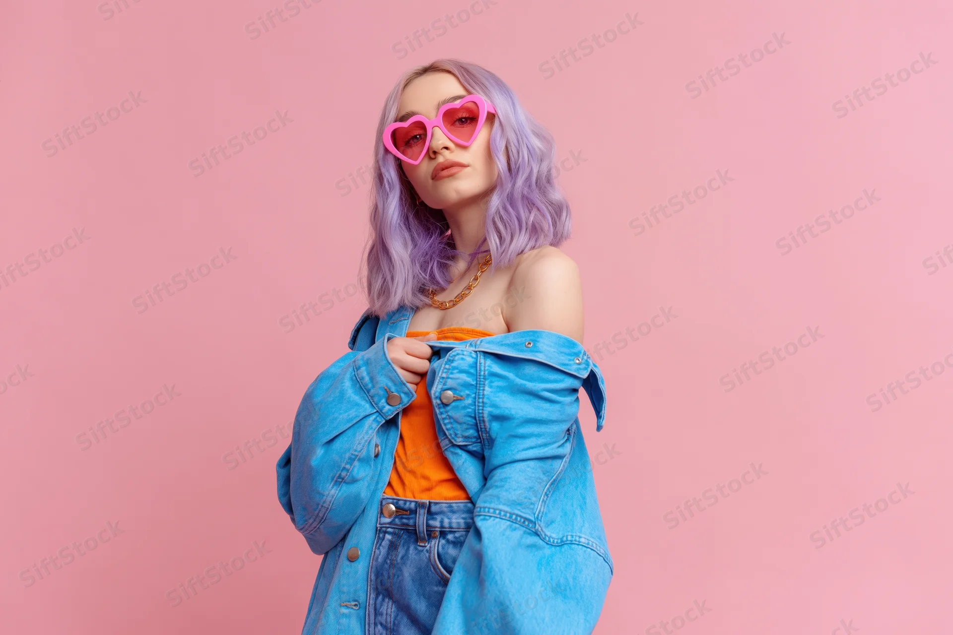 Young woman with lavender hair wearing heart-shaped sunglasses and denim jacket posing against pink background