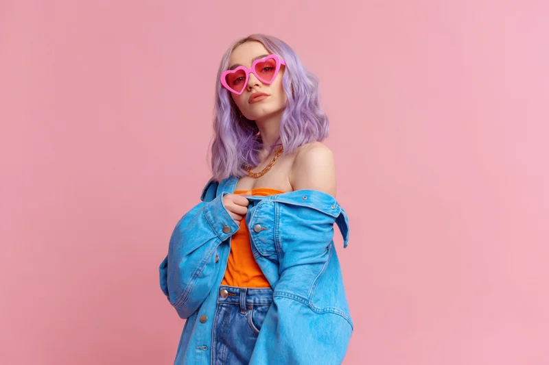 Young woman with lavender hair wearing heart-shaped sunglasses and denim jacket posing against pink background