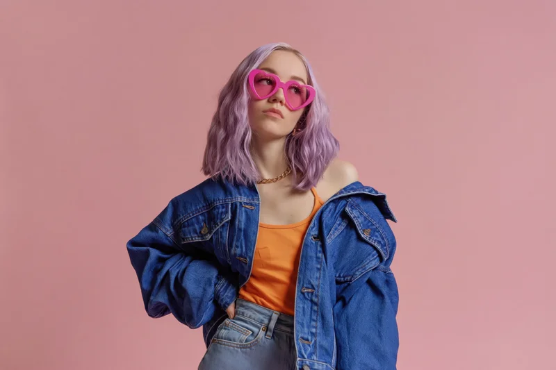 Young woman with lavender wavy hair wearing heart-shaped pink sunglasses and denim jacket posing in studio