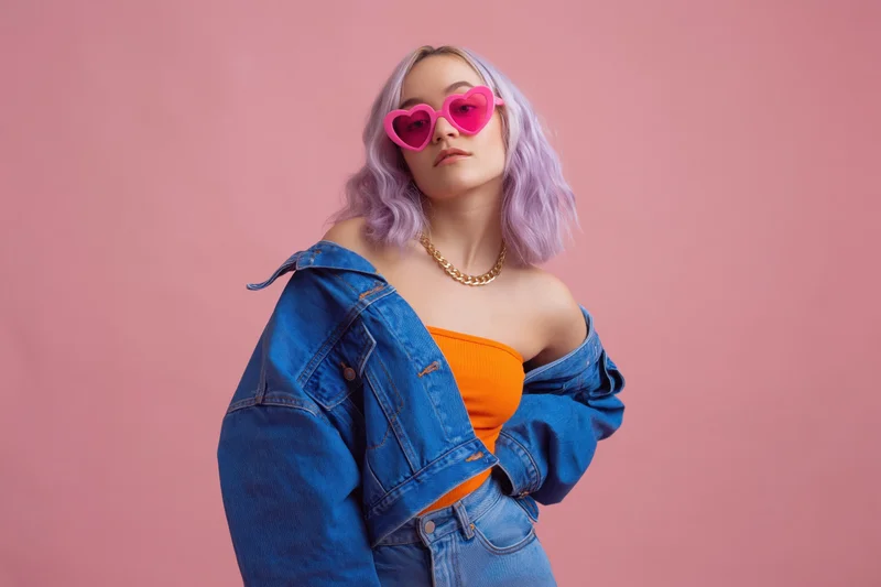Young woman with lavender hair wearing heart-shaped sunglasses and denim jacket posing in studio against pink background