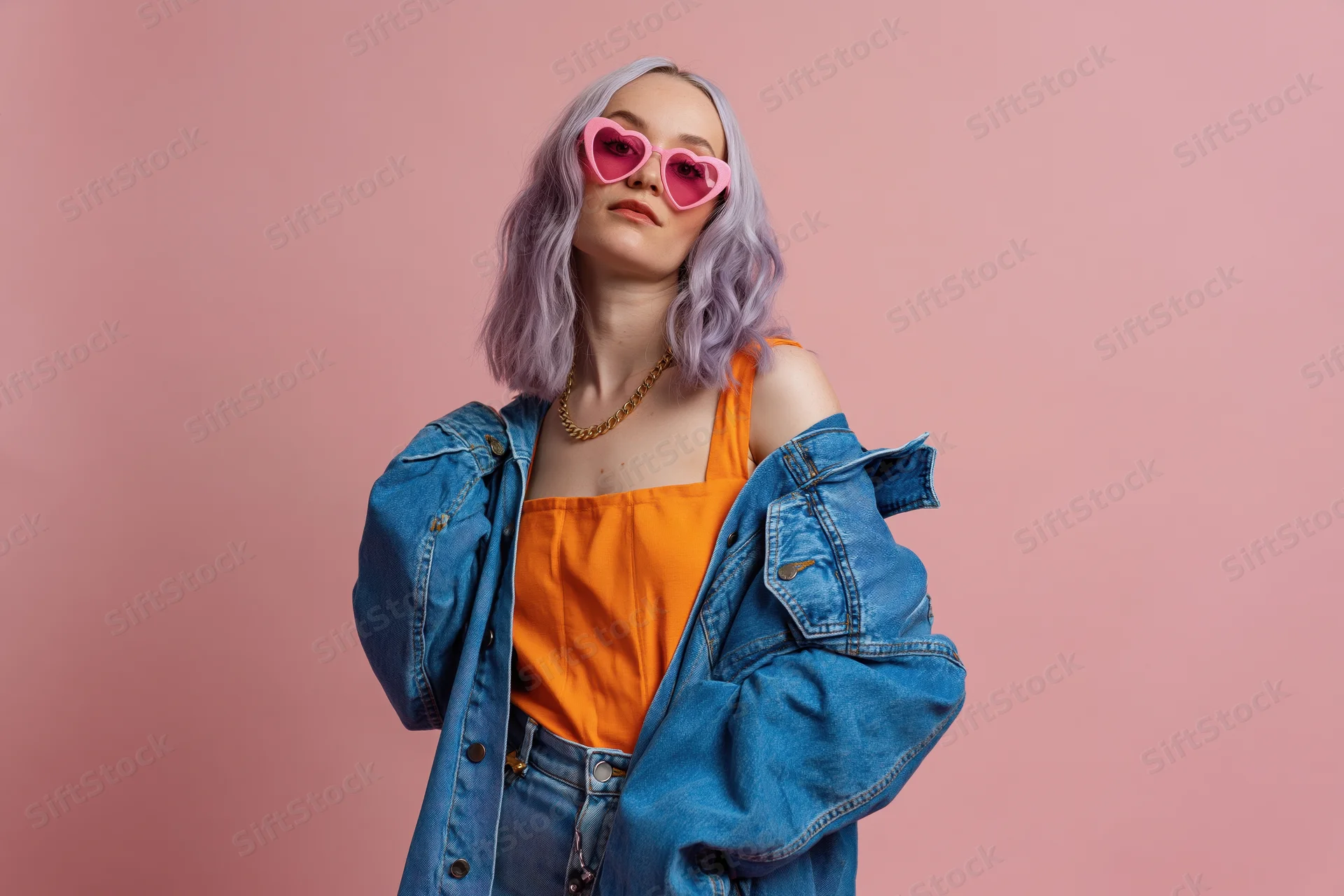 Young woman with lavender hair wearing heart-shaped sunglasses and denim jacket over orange top, posing against pink background