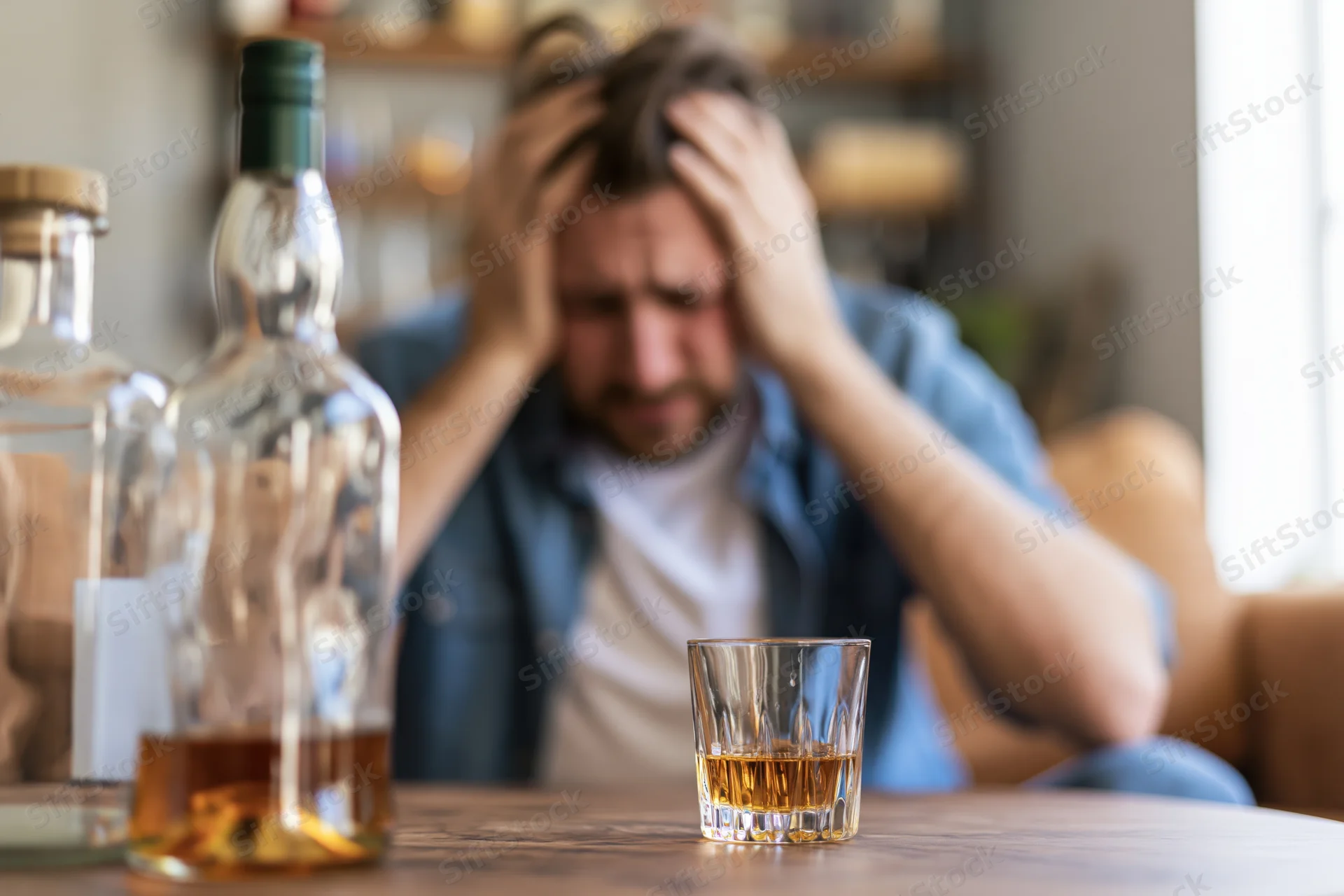 Distressed man holding head while sitting on sofa with whiskey glass and bottle on wooden table in living room