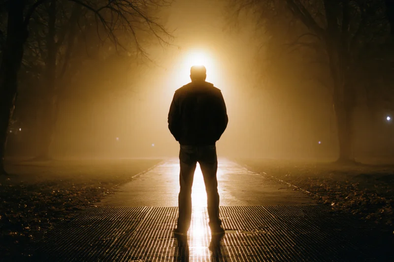 Man standing on wet metal grate path at night facing a glowing streetlight in foggy park