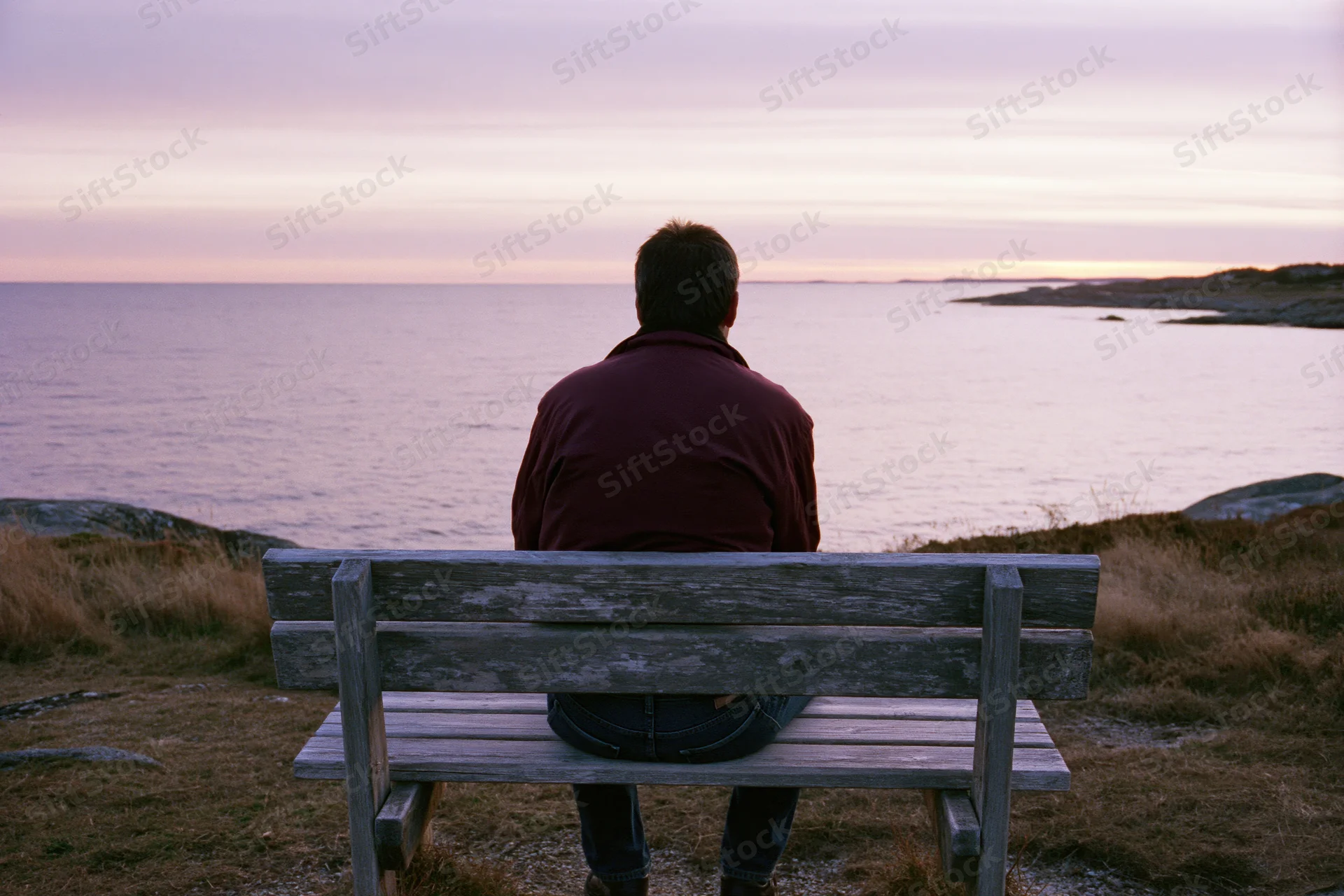 Man sitting alone on wooden bench facing ocean at sunset