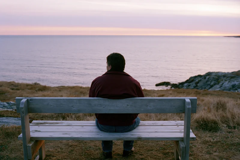 Man sitting alone on wooden bench facing ocean at sunset