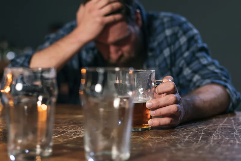Man slumped at wooden bar table gripping half-empty beer glass with hand on head