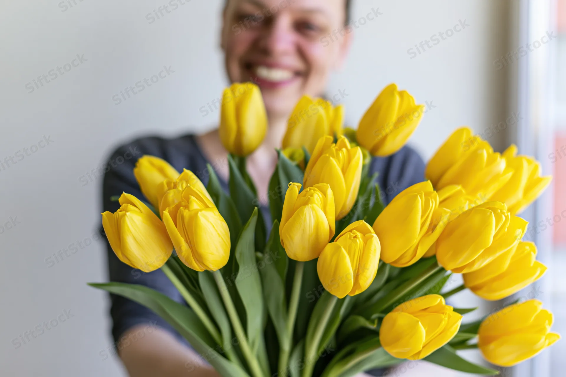 Woman smiling while holding a large bouquet of yellow tulips indoors near a window