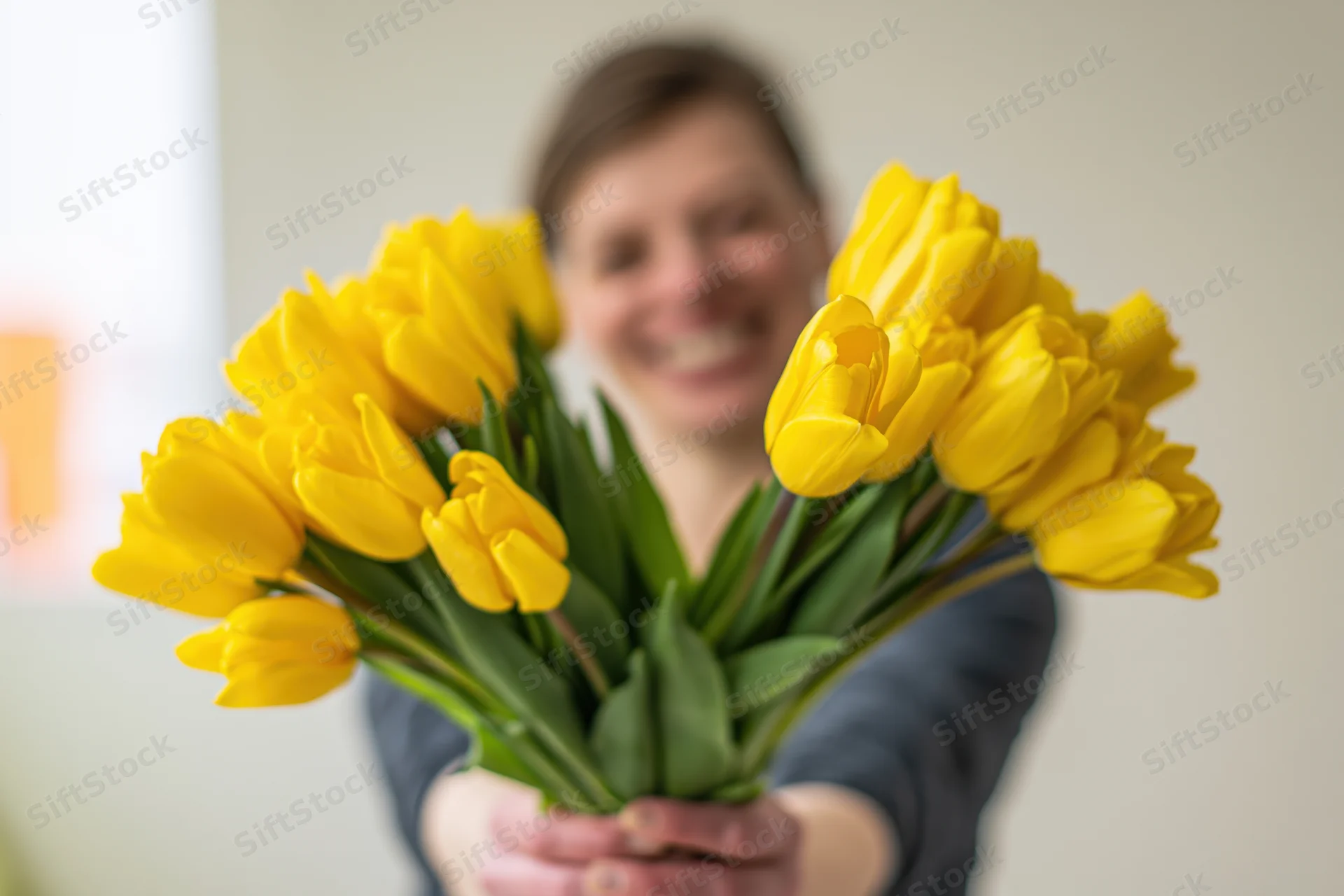 Woman holding out a bouquet of yellow tulips indoors, smiling in the background